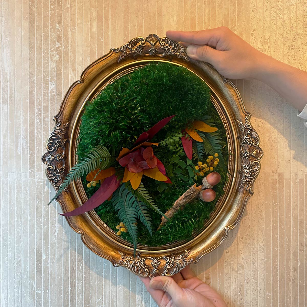 Handcrafted vintage oval framed moss wall art with autumn leaves, preserved greenery, and decorative acorns, shown held against a textured wall.